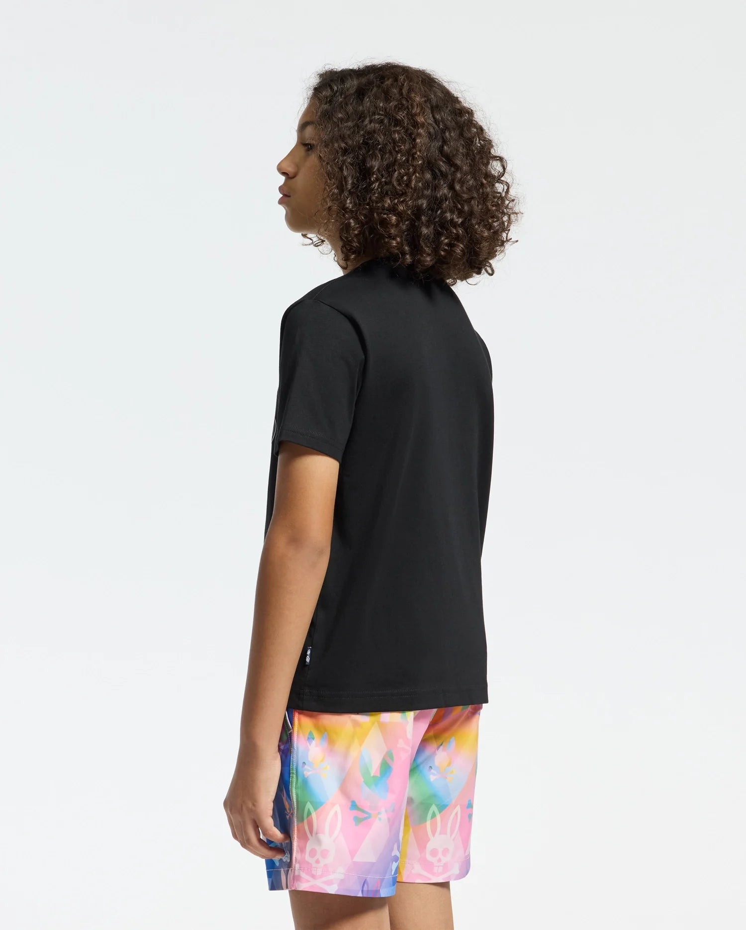 A person with curly hair, turned to the side, is wearing a Psycho Bunny KIDS MILOS GRAPHIC TEE (B0U456F200) featuring a center bunny graphic. Their vibrant shorts with playful patterns are made of soft Peruvian Pima cotton, all set against a plain white background.