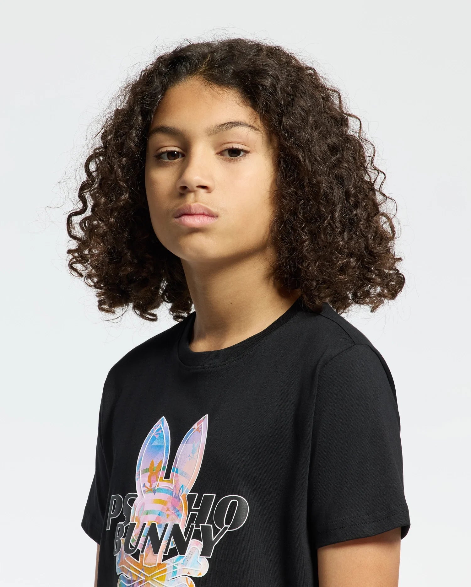 A curly-haired child wears a black "KIDS MILOS GRAPHIC TEE - B0U456F200" made of Peruvian Pima cotton, featuring a vivid bunny graphic and the text "Psycho Bunny" on a white background.