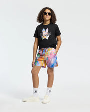 A person with curly hair and sunglasses confidently dons the Psycho Bunny KIDS MILOS GRAPHIC TEE - B0U456F200 in black, paired with colorful abstract shorts. The Peruvian Pima cotton attire is finished with white socks and sneakers, set against a plain white backdrop.