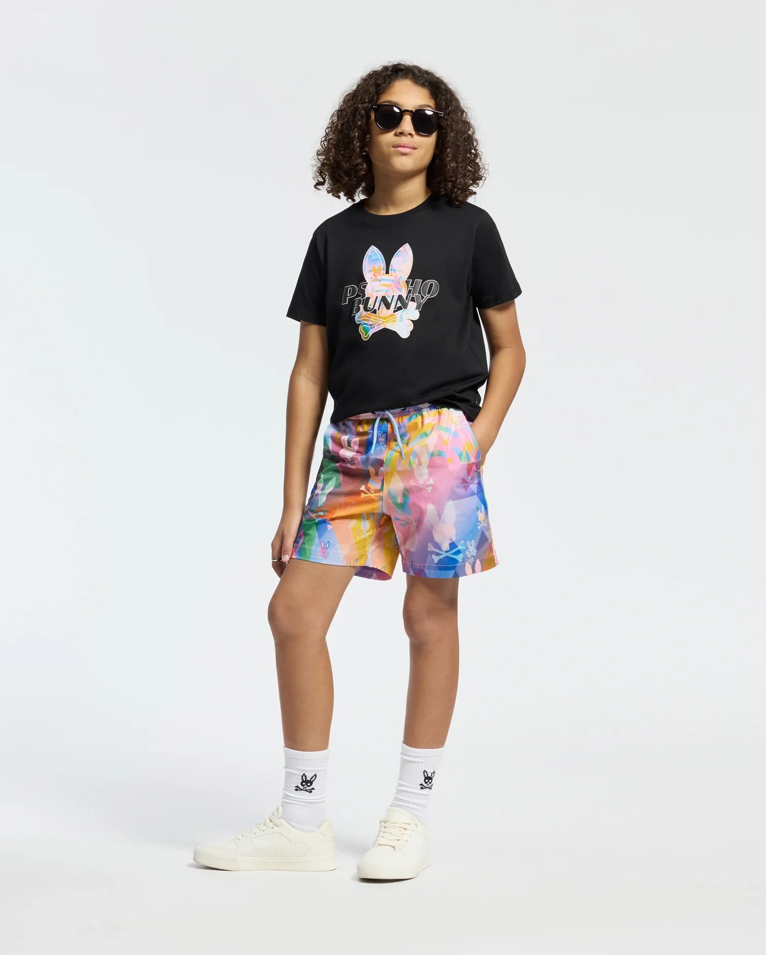A person with curly hair and sunglasses confidently dons the Psycho Bunny KIDS MILOS GRAPHIC TEE - B0U456F200 in black, paired with colorful abstract shorts. The Peruvian Pima cotton attire is finished with white socks and sneakers, set against a plain white backdrop.
