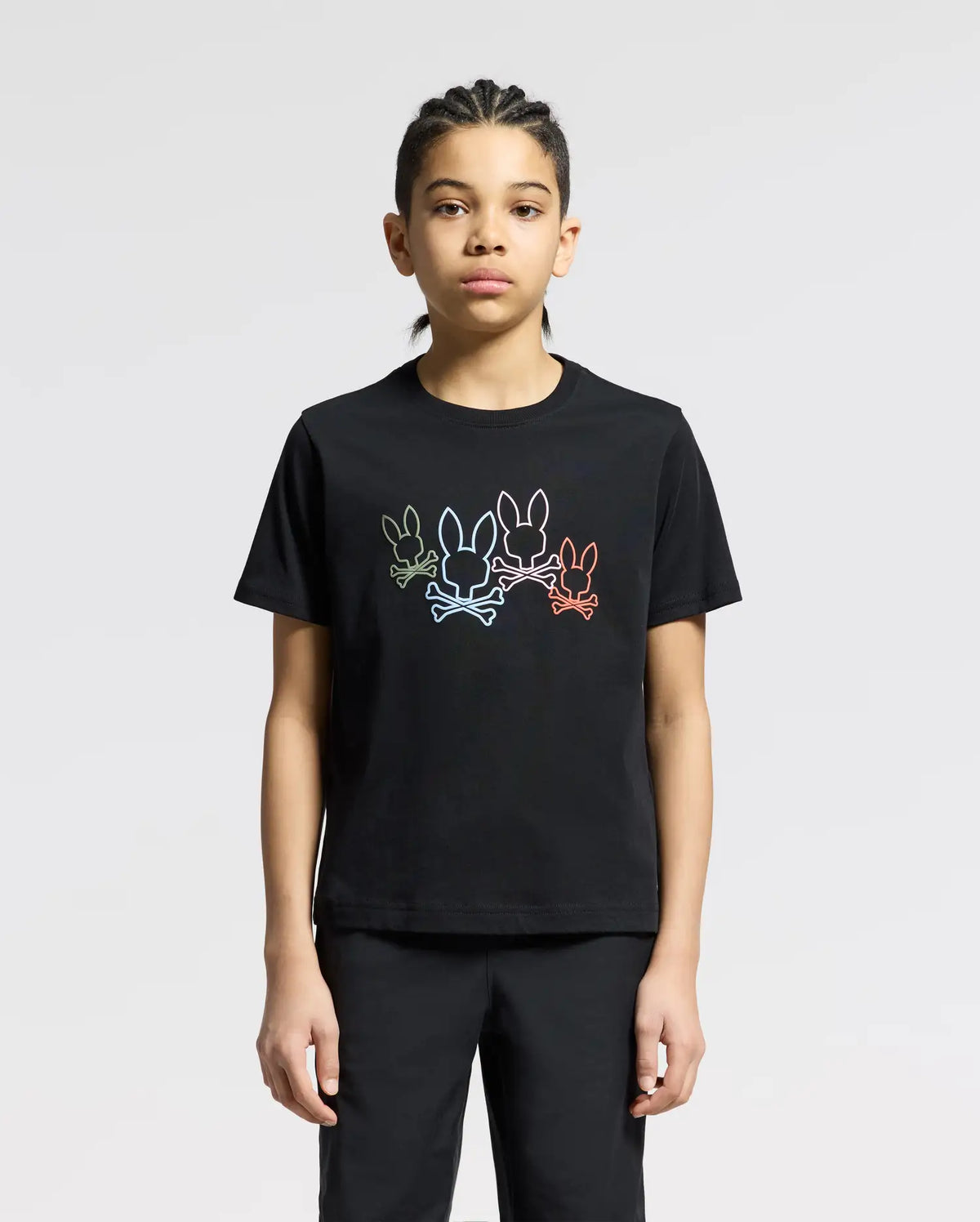 A person with braided hair wears a Psycho Bunny KIDS SILVERDALE GRAPHIC TEE - B0U499F200, made of soft Pima cotton and featuring a water-based print of outlined cartoon rabbits in various colors. They stand against a plain, light gray background.