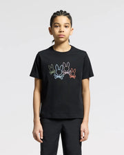 A person with braided hair wears a Psycho Bunny KIDS SILVERDALE GRAPHIC TEE - B0U499F200, made of soft Pima cotton and featuring a water-based print of outlined cartoon rabbits in various colors. They stand against a plain, light gray background.