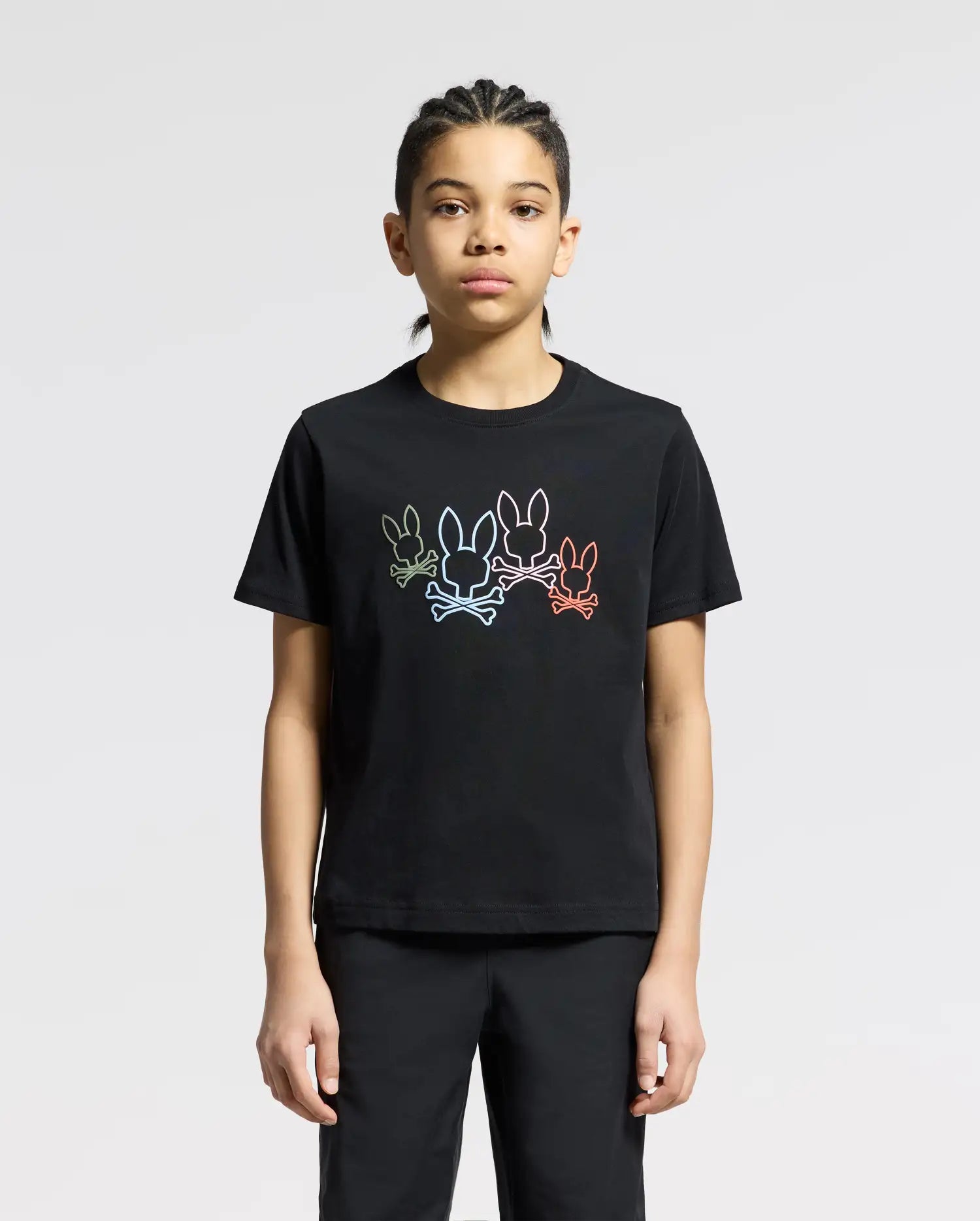 A person with braided hair wears a Psycho Bunny KIDS SILVERDALE GRAPHIC TEE - B0U499F200, made of soft Pima cotton and featuring a water-based print of outlined cartoon rabbits in various colors. They stand against a plain, light gray background.