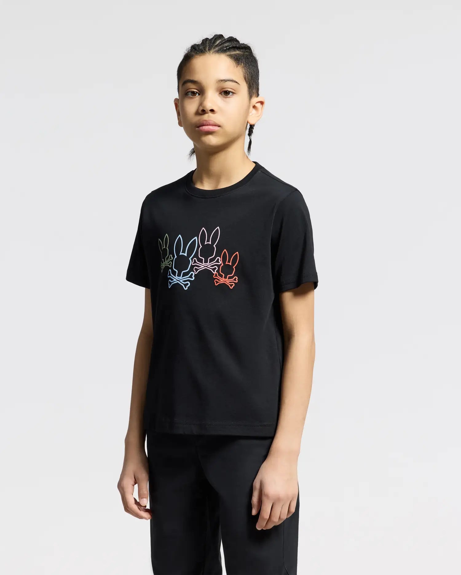 A person with braided hair stands against a plain background, wearing the Psycho Bunny KIDS SILVERDALE GRAPHIC TEE - B0U499F200 in black. The Pima cotton shirt features a water-based multicolor rabbit print. Their expression is neutral, and their hands are at their sides.
