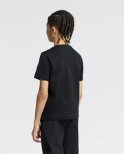 A person with intricately braided hair stands facing away, dressed in a black Psycho Bunny KIDS SILVERDALE GRAPHIC TEE - B0U499F200 featuring a subtle water-based Bunny print, paired with pants. The soft Pima cotton outfit is set against a plain white background.