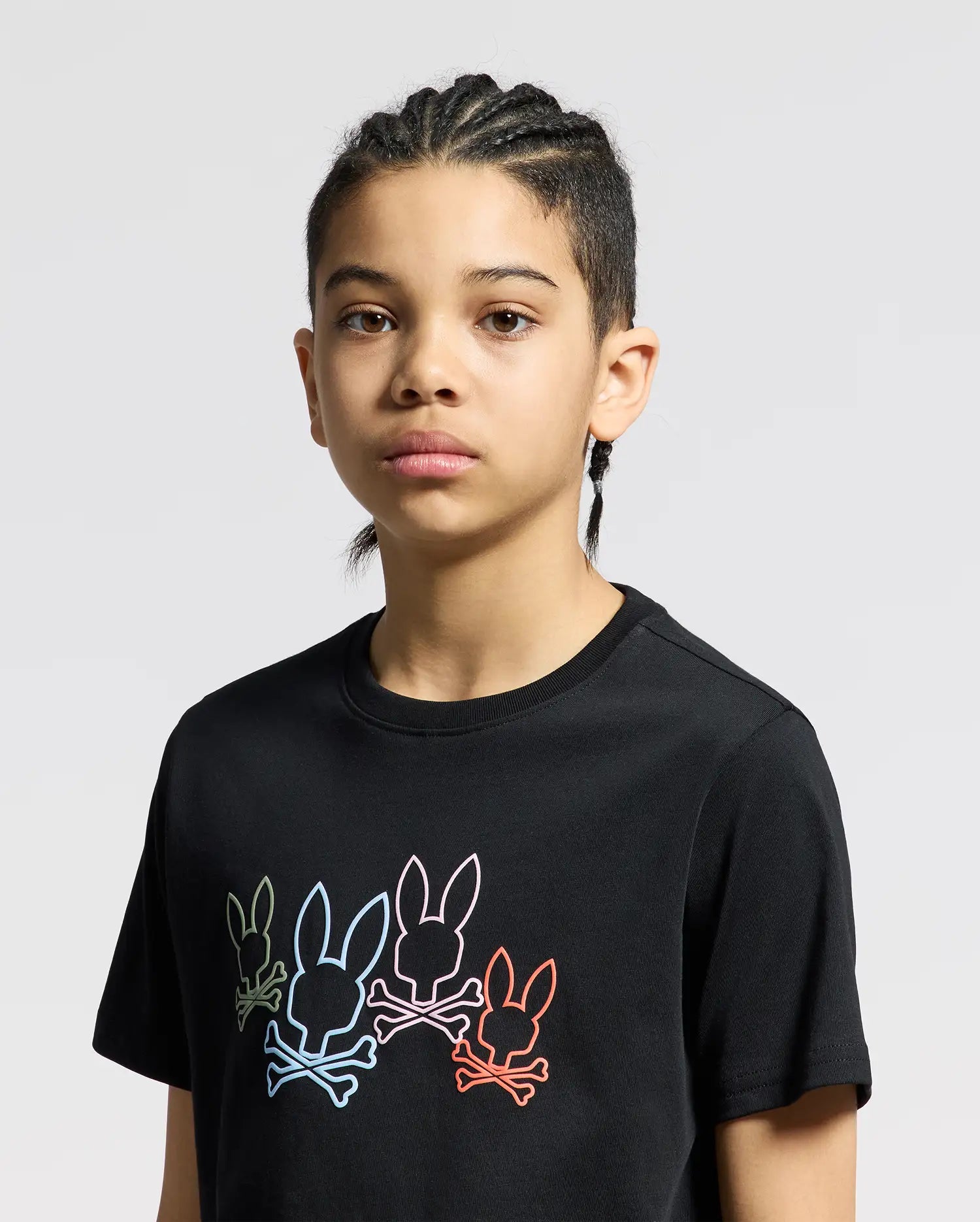 A person with braided hair wears the Psycho Bunny KIDS SILVERDALE GRAPHIC TEE featuring a vibrant bunny skull design on a black Pima cotton fabric, standing out against the plain gray background.