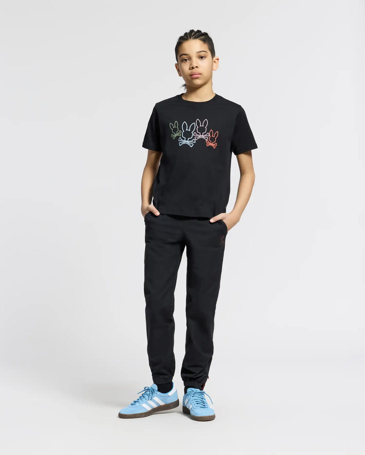 A young person confidently wears a KIDS SILVERDALE GRAPHIC TEE by Psycho Bunny, featuring a colorful bunny graphic. They're paired with matching black pants and light blue Adidas sneakers, hands in pockets against a plain light gray background.