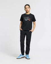 A young person confidently wears a KIDS SILVERDALE GRAPHIC TEE by Psycho Bunny, featuring a colorful bunny graphic. They're paired with matching black pants and light blue Adidas sneakers, hands in pockets against a plain light gray background.