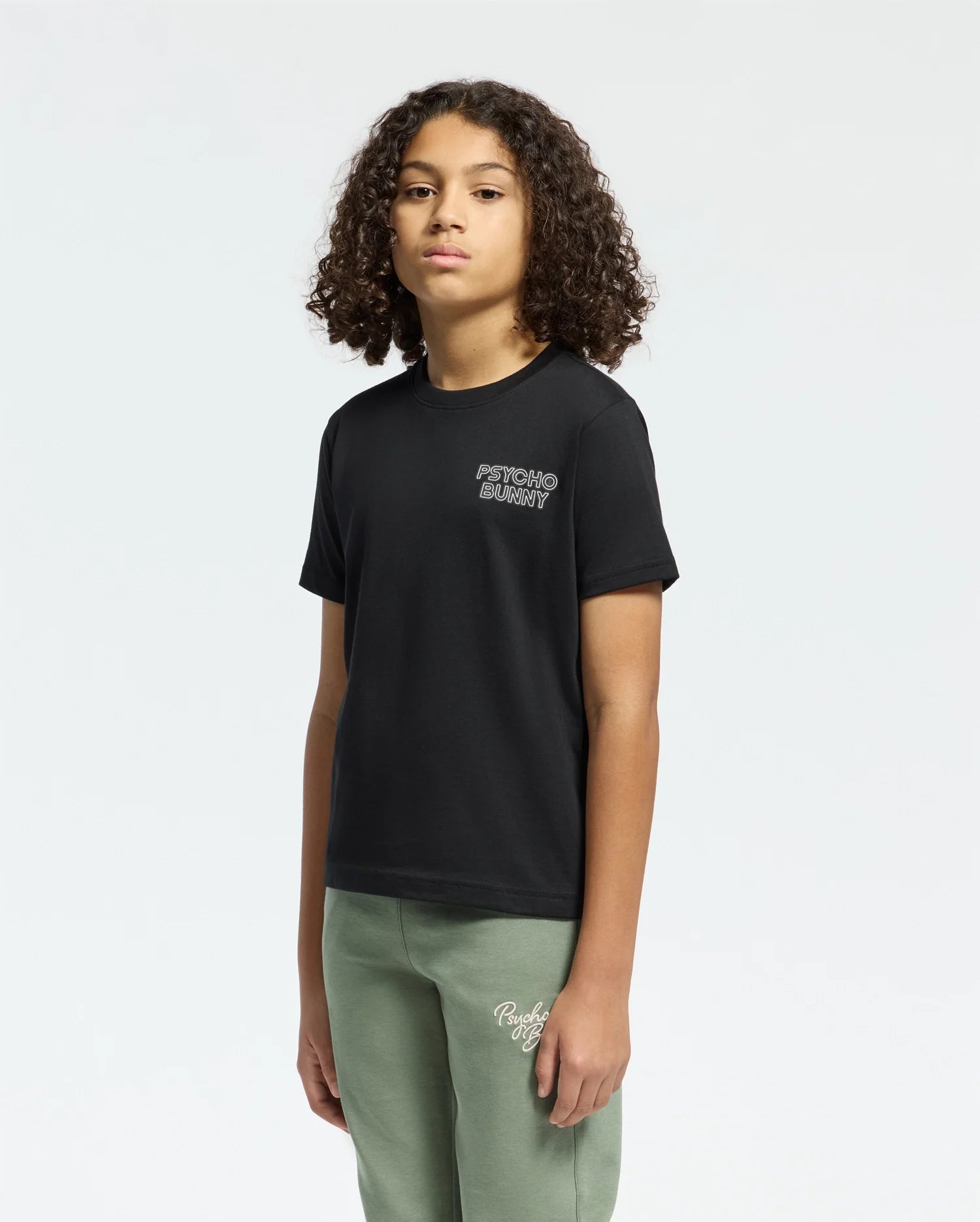 Against a plain backdrop, a person with curly hair sports the Psycho Bunny KIDS SYDNEY BACK GRAPHIC TEE - B0U509F200 in black Pima cotton, featuring a water-based print with white chest text and regular fit, paired with green pants. With hands at their sides, they have a neutral expression.