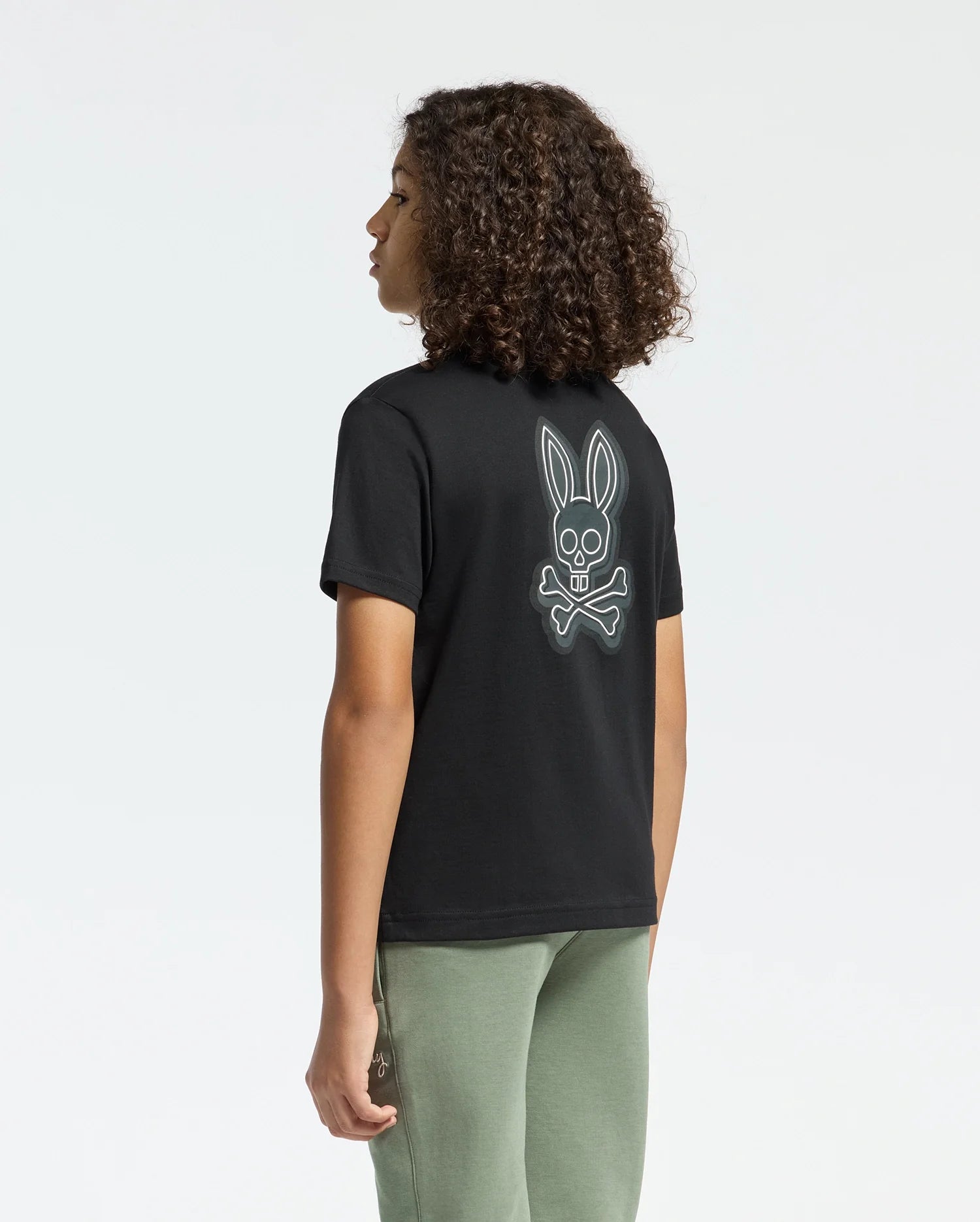 A curly-haired person wears the Psycho Bunny KIDS SYDNEY BACK GRAPHIC TEE (B0U509F200). This regular-fit, black Pima cotton T-shirt features a water-based print of a cartoon skull and crossbones with bunny ears on the back. They're paired with green pants against a plain white background.