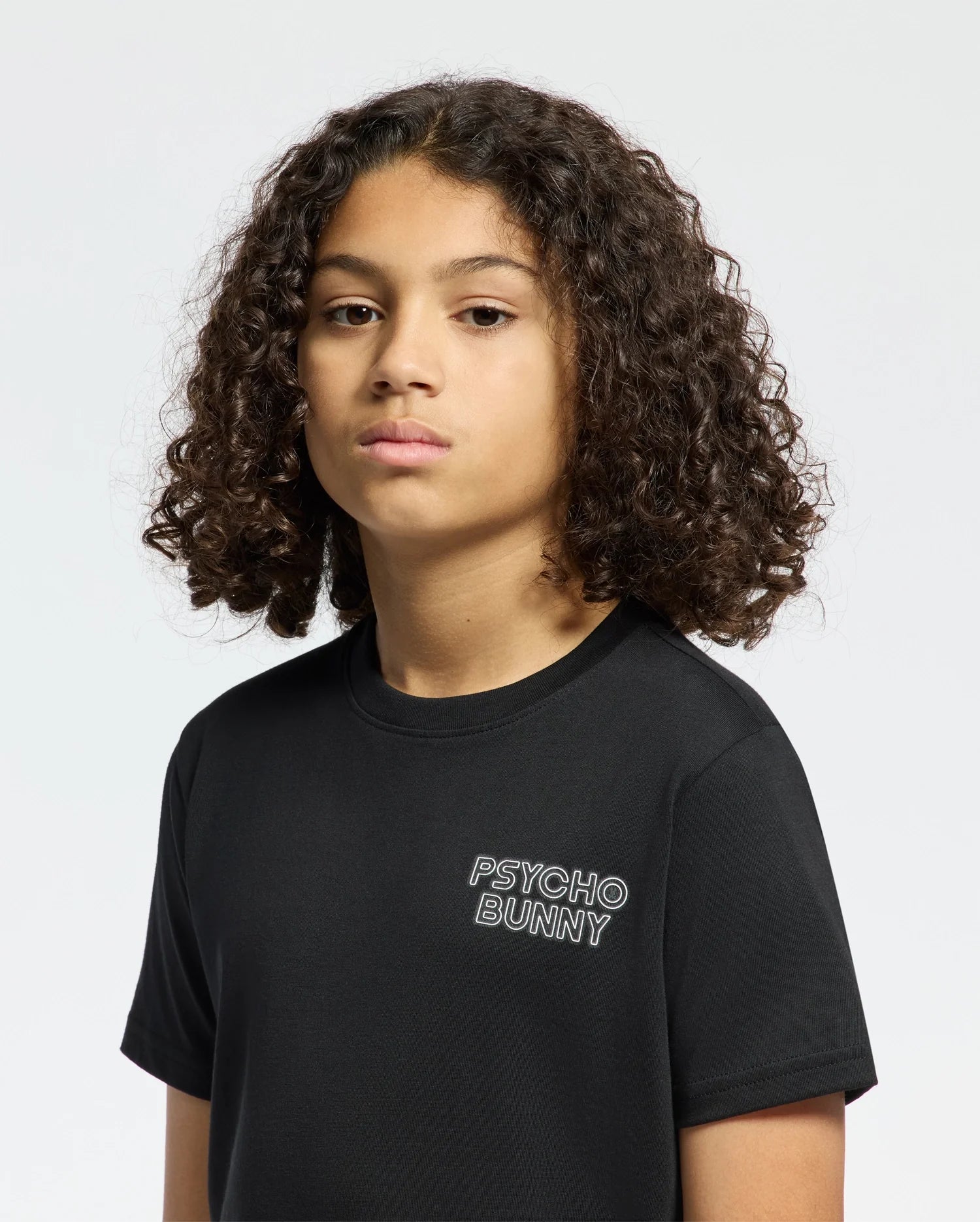 A curly-haired person wears the Psycho Bunny Kids Sydney Back Graphic Tee (B0U509F200), a regular fit black Pima cotton t-shirt, against a plain white background.