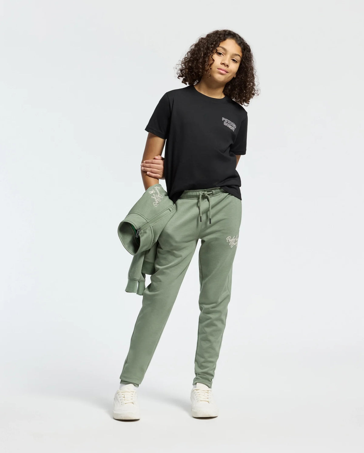 Against a plain background, a curly-haired person wears a black KIDS SYDNEY BACK GRAPHIC TEE by Psycho Bunny and green Pima cotton sweatpants, holding a matching green sweatshirt. They complete their relaxed look with white sneakers.