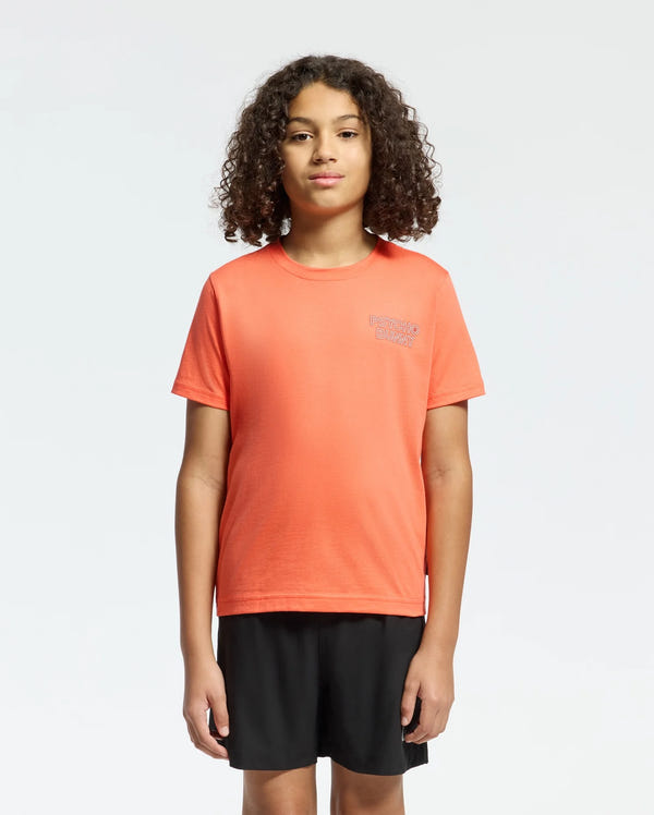 Against a plain background, a person with curly hair wears the KIDS SYDNEY BACK GRAPHIC TEE by Psycho Bunny in a regular fit, orange Pima cotton. The chest has small white water-based print text. They pair it with black shorts and gaze forward neutrally.