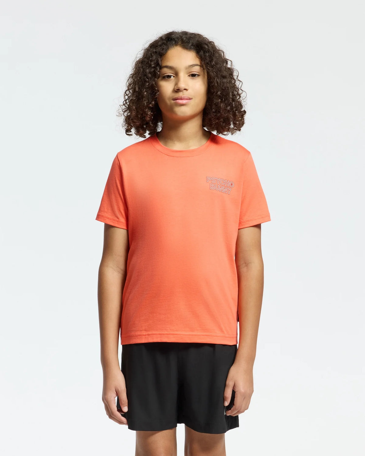 Against a plain background, a person with curly hair wears the KIDS SYDNEY BACK GRAPHIC TEE by Psycho Bunny in a regular fit, orange Pima cotton. The chest has small white water-based print text. They pair it with black shorts and gaze forward neutrally.