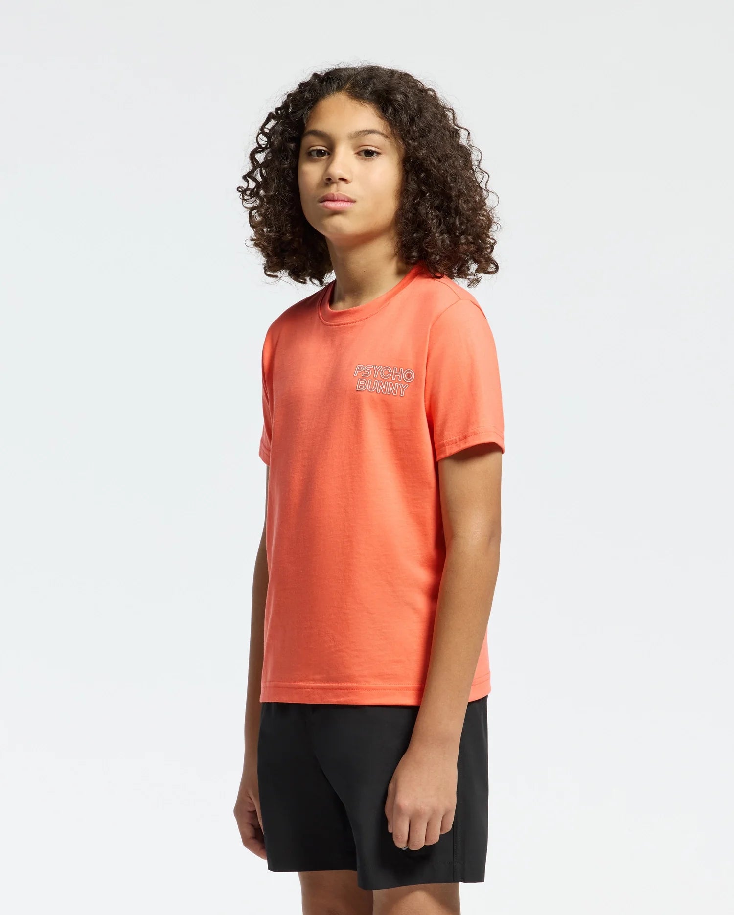A person with curly hair stands slightly sideways, dressed in an orange Psycho Bunny Kids Sydney Back Graphic Tee and black shorts. The Pima cotton shirt has a water-based print logo on the chest. The background is plain white.