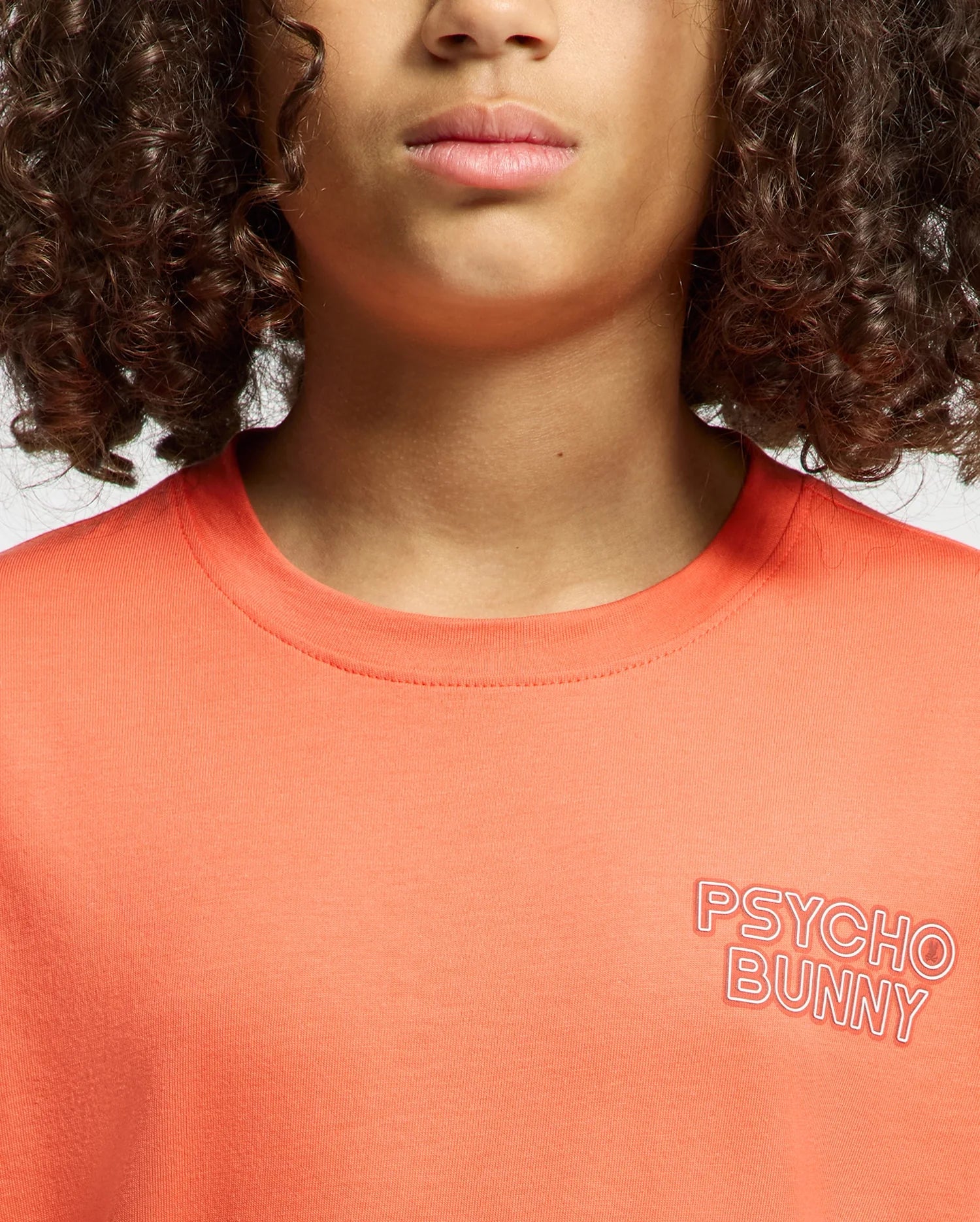 A person with curly hair wears the regular-fit orange "KIDS SYDNEY BACK GRAPHIC TEE" made of Pima cotton. It features a "Psycho Bunny" water-based print on the chest. The image focuses on the shirt and part of their face, emphasizing their closed lips.