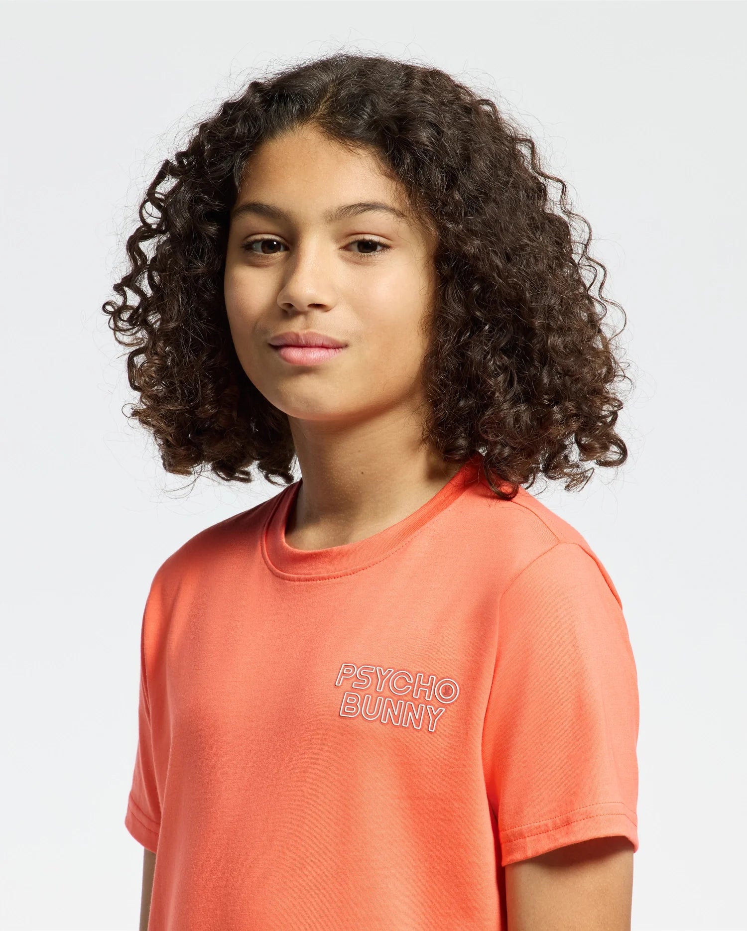 A young person with curly hair wears the Psycho Bunny KIDS SYDNEY BACK GRAPHIC TEE - B0U509F200 in orange, made from soft Pima cotton, featuring "Psycho Bunny" in a trendy water-based print, against a plain white background.