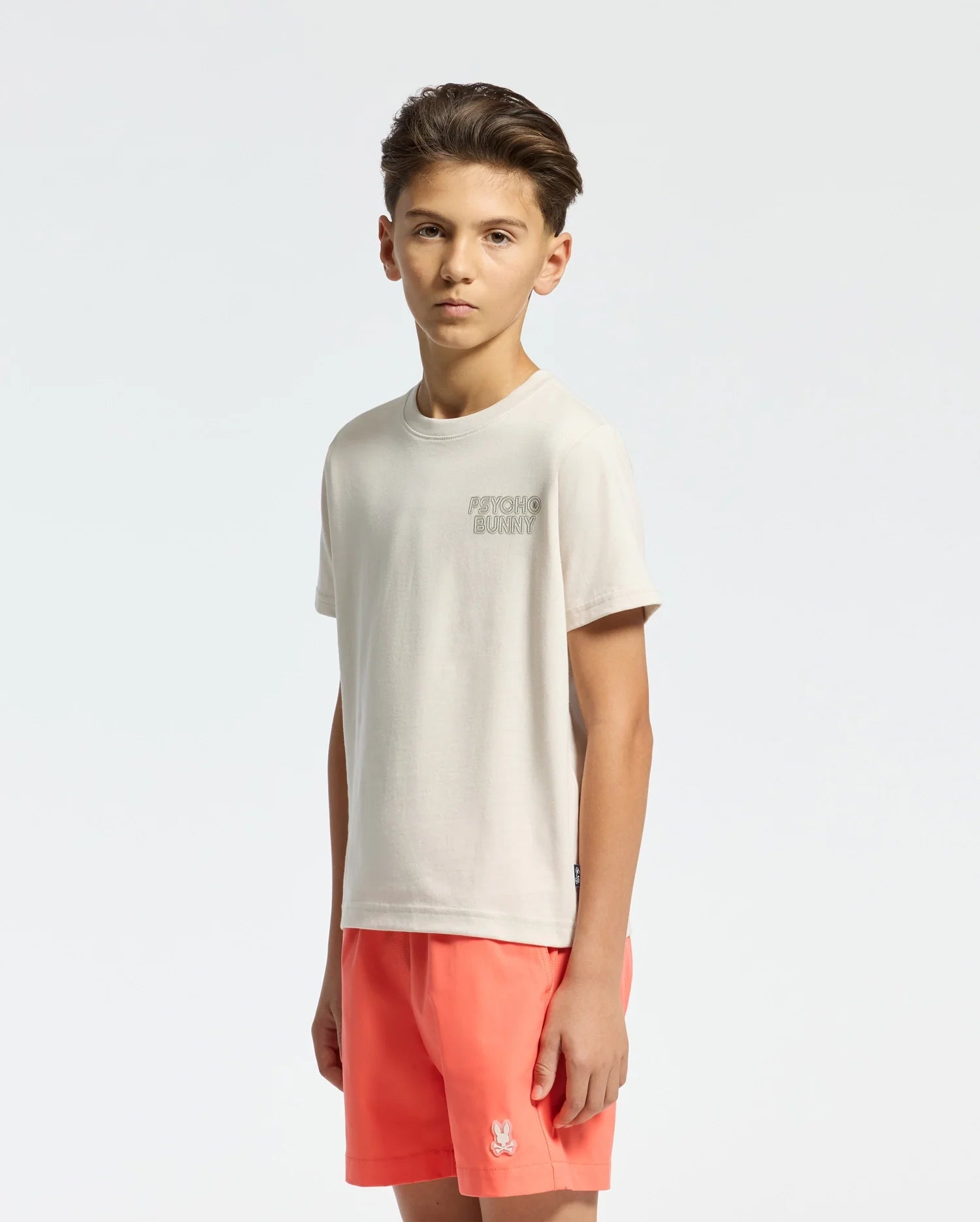 A young boy with short brown hair stands against a plain background, neutral-faced in a Psycho Bunny KIDS SYDNEY BACK GRAPHIC TEE - B0U509F200, made of light-colored Pima cotton, paired with bright coral shorts featuring a small water-based print emblem on the leg.