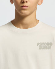 A child in a cream, regular-fit KIDS SYDNEY BACK GRAPHIC TEE (B0U509F200) by Psycho Bunny is shown. The tee, with "Psycho Bunny" embroidered on the upper left side, is the focus, along with their lower face in neutral expression against a plain background.