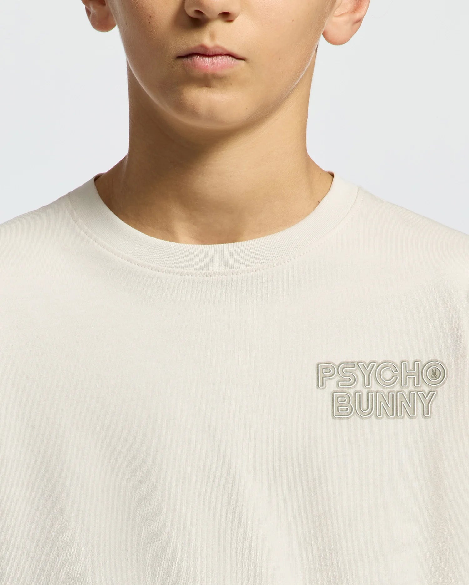 A child in a cream, regular-fit KIDS SYDNEY BACK GRAPHIC TEE (B0U509F200) by Psycho Bunny is shown. The tee, with "Psycho Bunny" embroidered on the upper left side, is the focus, along with their lower face in neutral expression against a plain background.