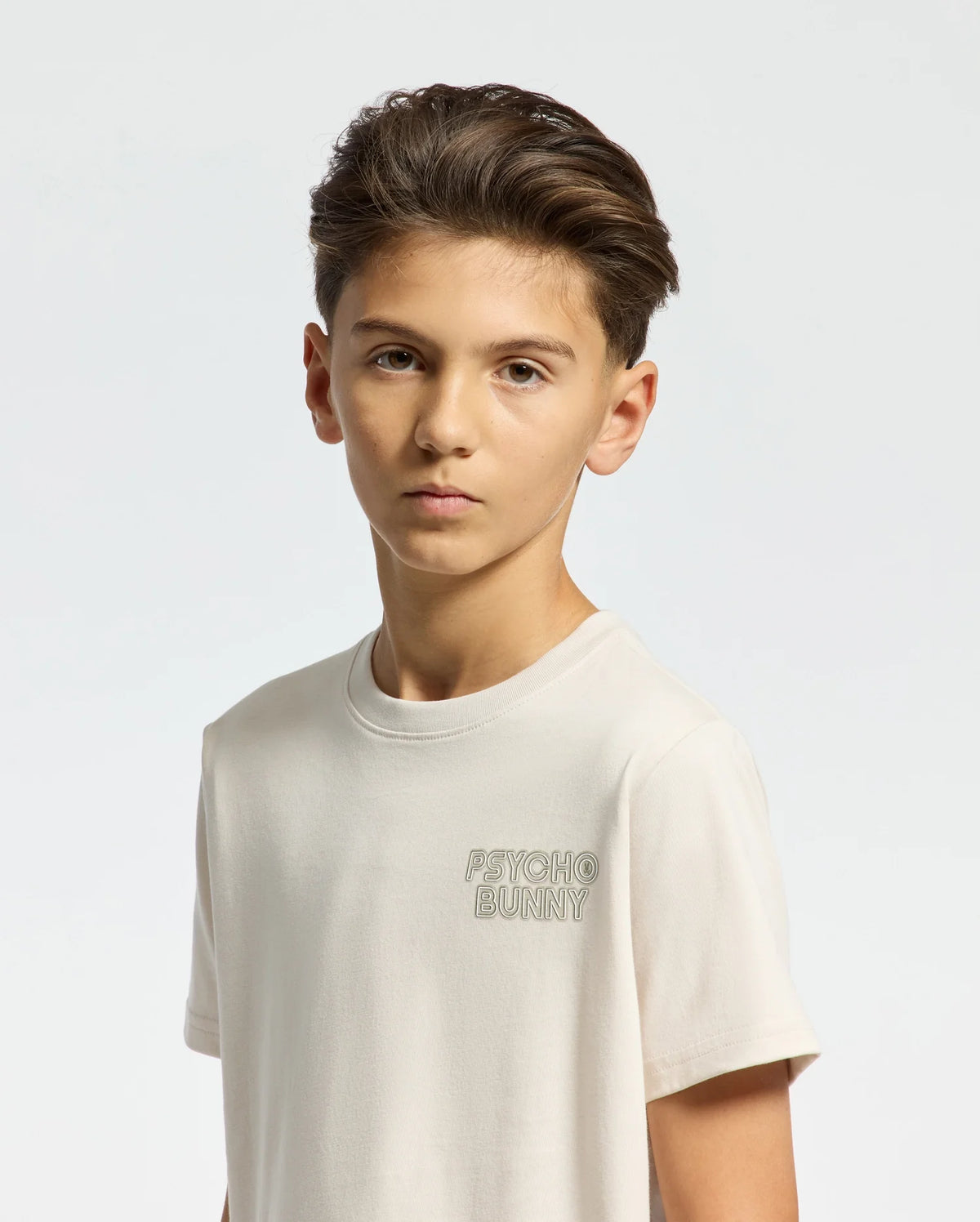 The boy with short brown hair sports a "KIDS SYDNEY BACK GRAPHIC TEE - B0U509F200" by Psycho Bunny, featuring a water-based logo print on a light t-shirt. The regular fit and Pima cotton material enhance his relaxed yet polished look against the plain background.
