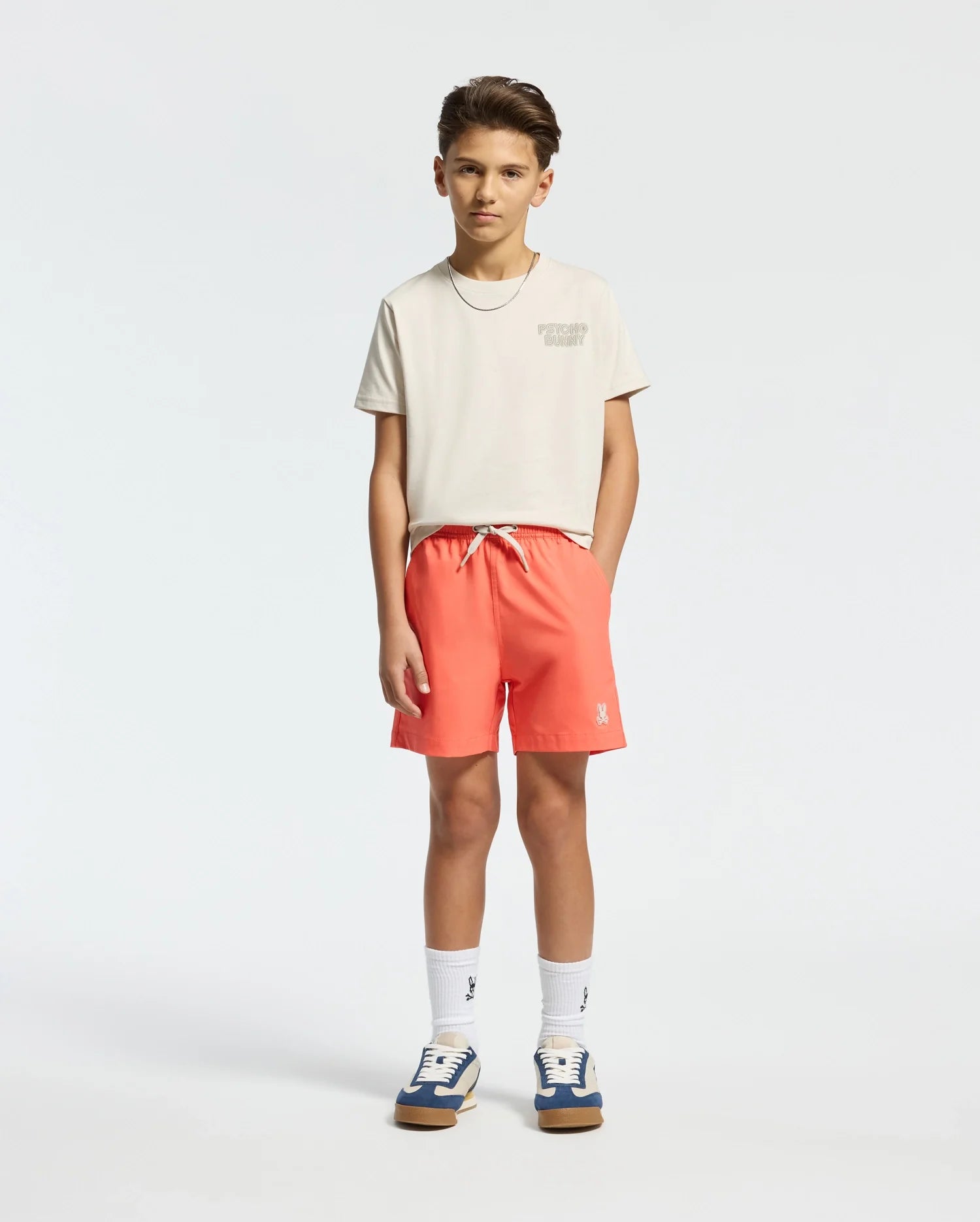 A young boy stands confidently, wearing a Psycho Bunny KIDS SYDNEY BACK GRAPHIC TEE in off-white and bright coral shorts, with white socks and sneakers featuring blue and beige accents. His hands are in his pockets as he gazes directly at the camera against a plain backdrop.