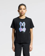 A youngster wears a Psycho Bunny KIDS ANDROS GRAPHIC TEE - B0U525F200, showcasing a neon-style bunny above crossbones. Made from 100% Pima Cotton in Peru, they stand against a light gray backdrop, gazing at the camera.