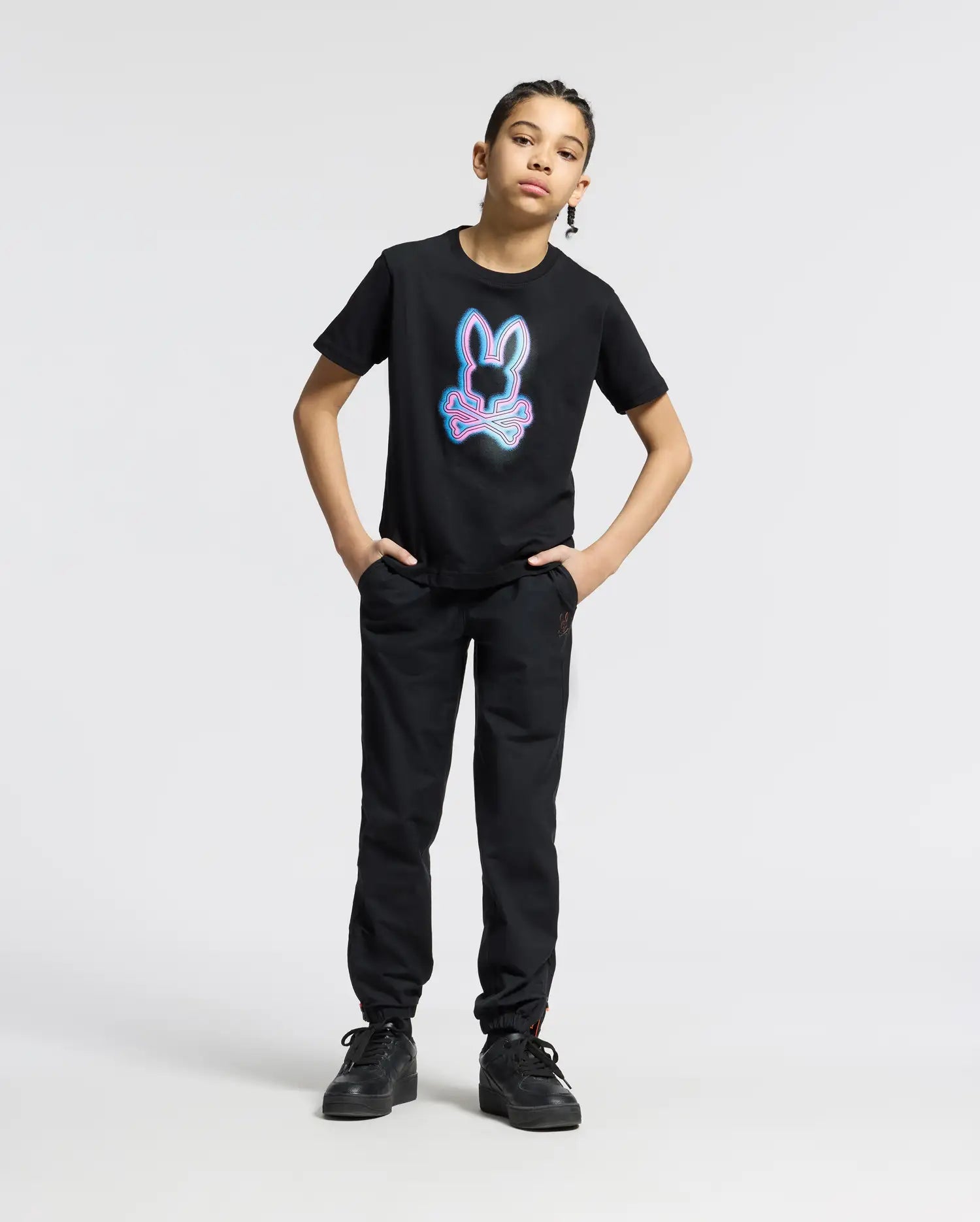 A child stands confidently with hands in pockets, wearing the Psycho Bunny KIDS ANDROS GRAPHIC TEE - B0U525F200. The 100% Pima Cotton black tee from Peru displays a colorful bunny design, highlighted against a plain white background.