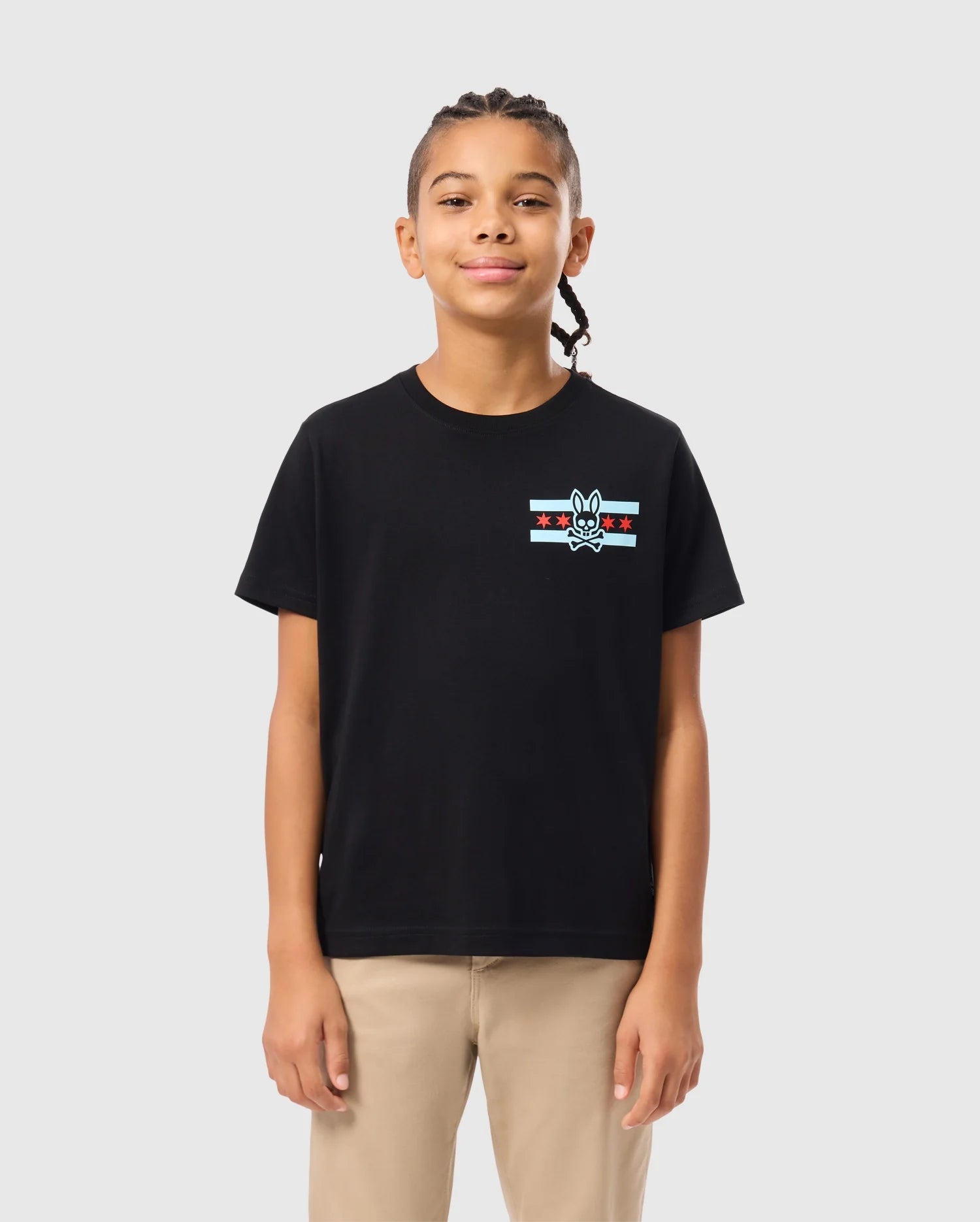 A young person with braided hair is wearing the KIDS CHICAGO CITY TEE from Psycho Bunny, a black Pima cotton jersey featuring red and white stripes and a small emblem, paired with beige pants, standing against a plain light gray background.