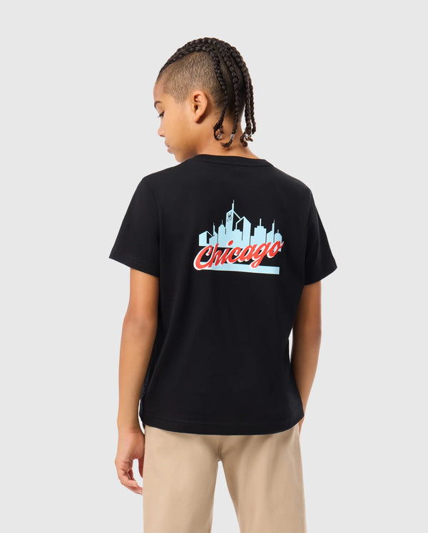 A child with braided hair stands facing away, wearing the Psycho Bunny KIDS CHICAGO CITY TEE - B0U551W1PC, a black t-shirt crafted from soft Pima cotton jersey and showcasing the word "Chicago" along with a skyline graphic. Light-colored pants complete this casual look.