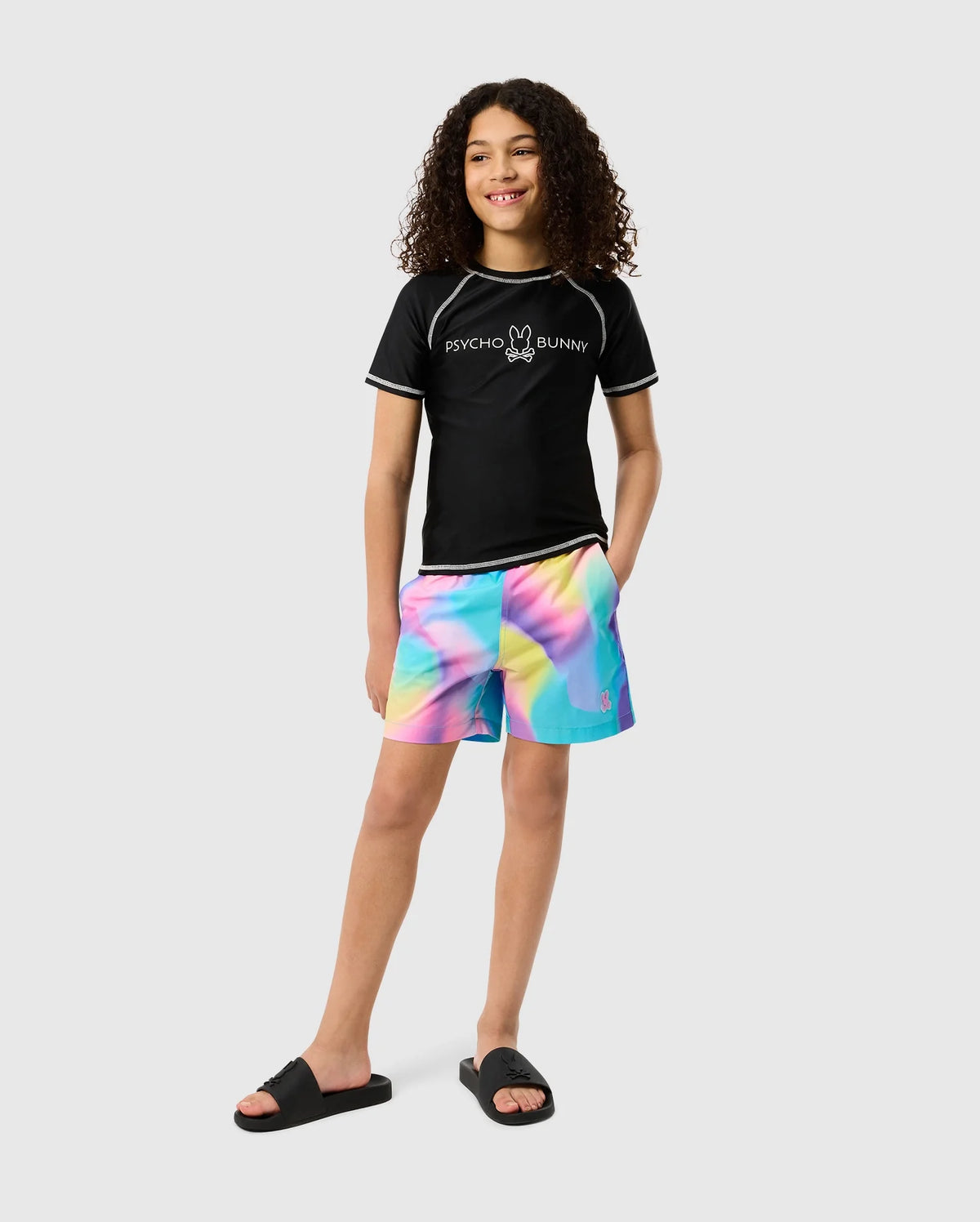 A person with long curly hair stands confidently, wearing a black "Psycho Bunny" T-shirt, colorful tie-dye shorts, and black sandals. The KIDS SHORT SLEEVE ANTI-UV RASHGUARD - B0U558C200 by Psycho Bunny provides UPF 50+ protection against the sun's rays, making it perfect for a stylish yet safe day out. The background is plain white.