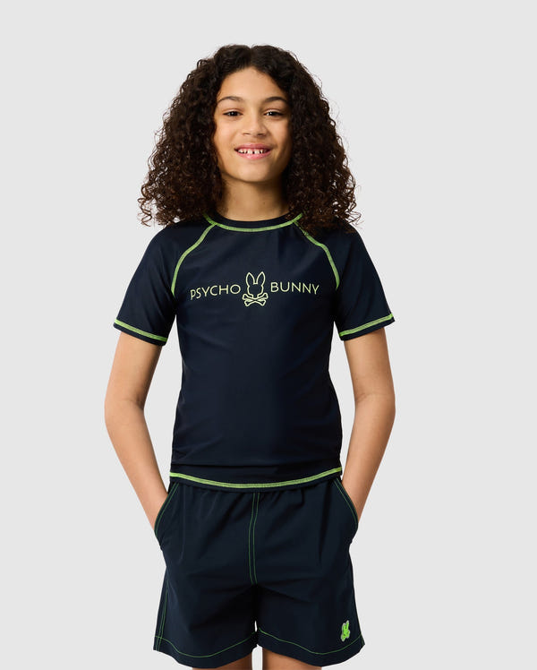 A young person with curly hair wears a navy KIDS SHORT SLEEVE RASH GUARD featuring neon green stitching and the Psycho Bunny logo. Their hands are in their shorts pockets as they smile against a plain backdrop.