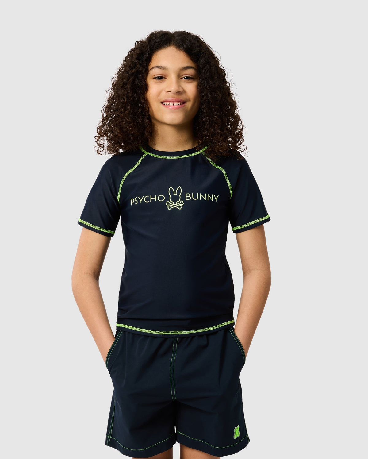 A young person with curly hair wears a navy KIDS SHORT SLEEVE RASH GUARD featuring neon green stitching and the Psycho Bunny logo. Their hands are in their shorts pockets as they smile against a plain backdrop.