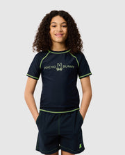 A young person with curly hair wears a navy KIDS SHORT SLEEVE RASH GUARD featuring neon green stitching and the Psycho Bunny logo. Their hands are in their shorts pockets as they smile against a plain backdrop.