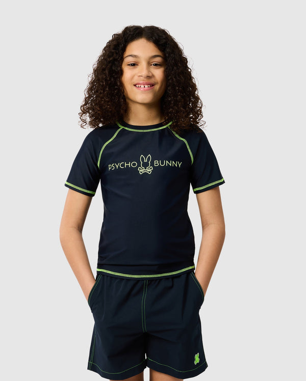 A young person with curly hair wears a navy KIDS SHORT SLEEVE RASH GUARD featuring neon green stitching and the Psycho Bunny logo. Their hands are in their shorts pockets as they smile against a plain backdrop.