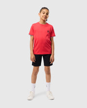 A child stands against a plain background wearing a red short-sleeved KIDS KAYDEN FASHION TEE - B0U577C200 by Psycho Bunny with black trim on the sleeves and a small embroidered chest Bunny on the left chest. They are also wearing black shorts, white socks, and white sneakers. They are smiling with their hands at their sides.