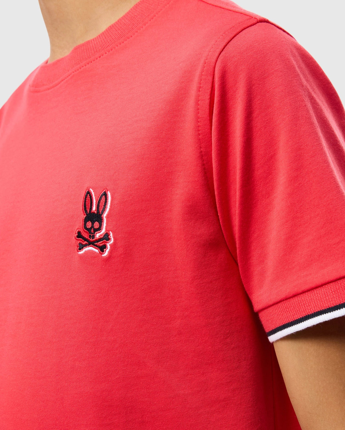 Close-up of a person wearing a red Psycho Bunny KIDS KAYDEN FASHION TEE - B0U577C200 with an embroidered chest Bunny skull and crossbones logo. The sleeve has a white, black, and red striped cuff detail. Made from soft Pima cotton, the background is plain and light-colored.