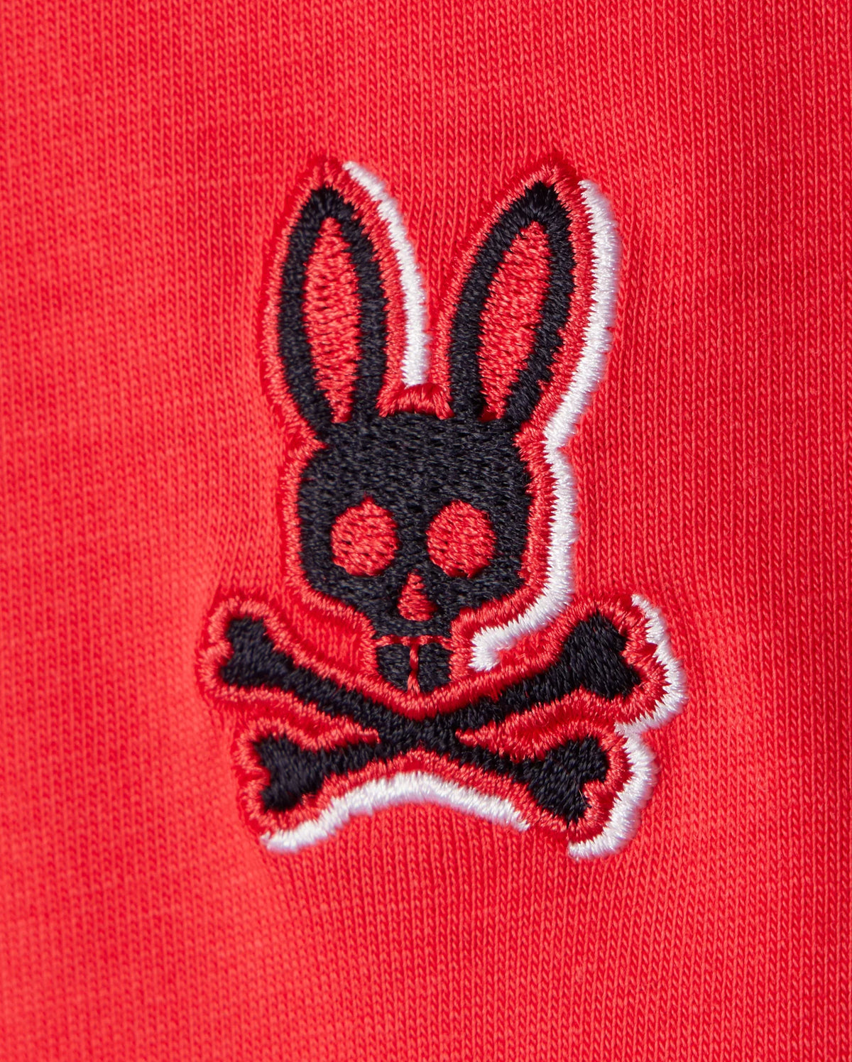 A close-up of an embroidered chest Bunny logo on red Pima cotton fabric. The logo features a black bunny head with long ears and large round eyes, positioned above a pair of crossed bones, reminiscent of a skull and crossbones design. The edges of the logo are trimmed in white, perfect for the KIDS KAYDEN FASHION TEE - B0U577C200 by Psycho Bunny.