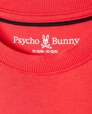 Close-up of the neck label on a red KIDS KAYDEN FASHION TEE - B0U577C200 by Psycho Bunny, featuring the brand's name and logo of a bunny skull with crossbones. The label indicates the size as 10-12/M. The stitching details of the collar and neckline are visible, highlighting the premium Pima cotton used for this stylish piece.