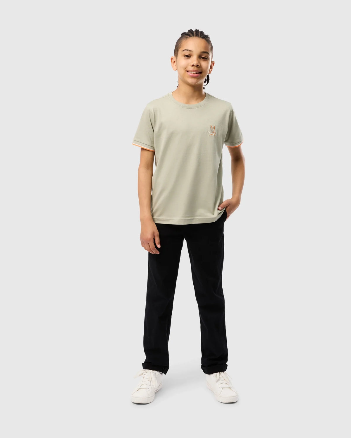 A young person with braided hair is standing against a plain background. They are wearing a light green KIDS KAYDEN FASHION TEE - B0U577C200 by Psycho Bunny, black pants, and white sneakers. They are smiling and have one hand in their pocket.