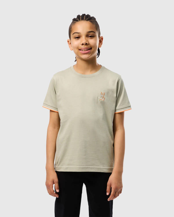 A smiling child with braided hair is standing against a plain background. They are wearing a light gray KIDS KAYDEN FASHION TEE - B0U577C200 by Psycho Bunny, made of soft Pima cotton, featuring an embroidered chest bunny on the left side, and black pants. The T-shirt has orange trim on the sleeves.