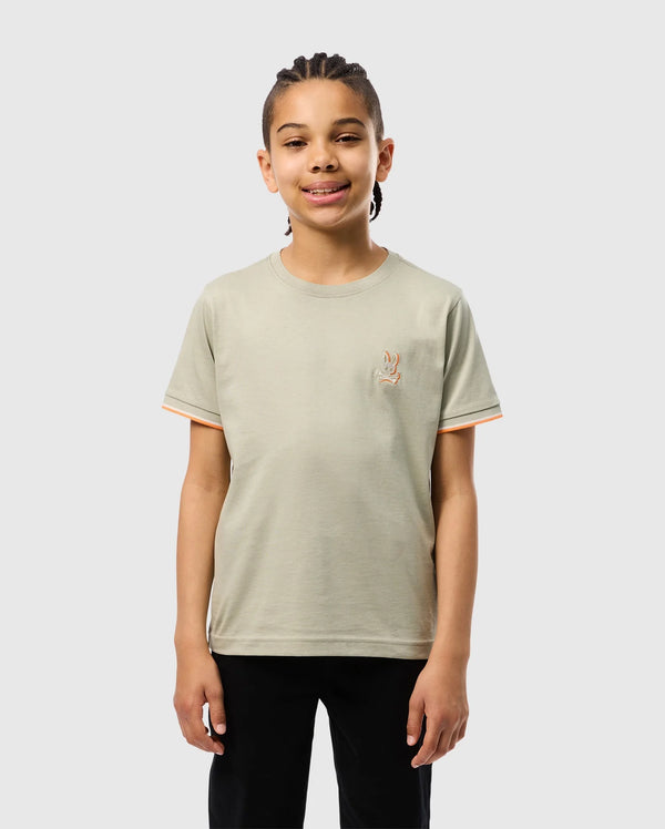 A smiling child with braided hair is standing against a plain background. They are wearing a light gray KIDS KAYDEN FASHION TEE - B0U577C200 by Psycho Bunny, made of soft Pima cotton, featuring an embroidered chest bunny on the left side, and black pants. The T-shirt has orange trim on the sleeves.