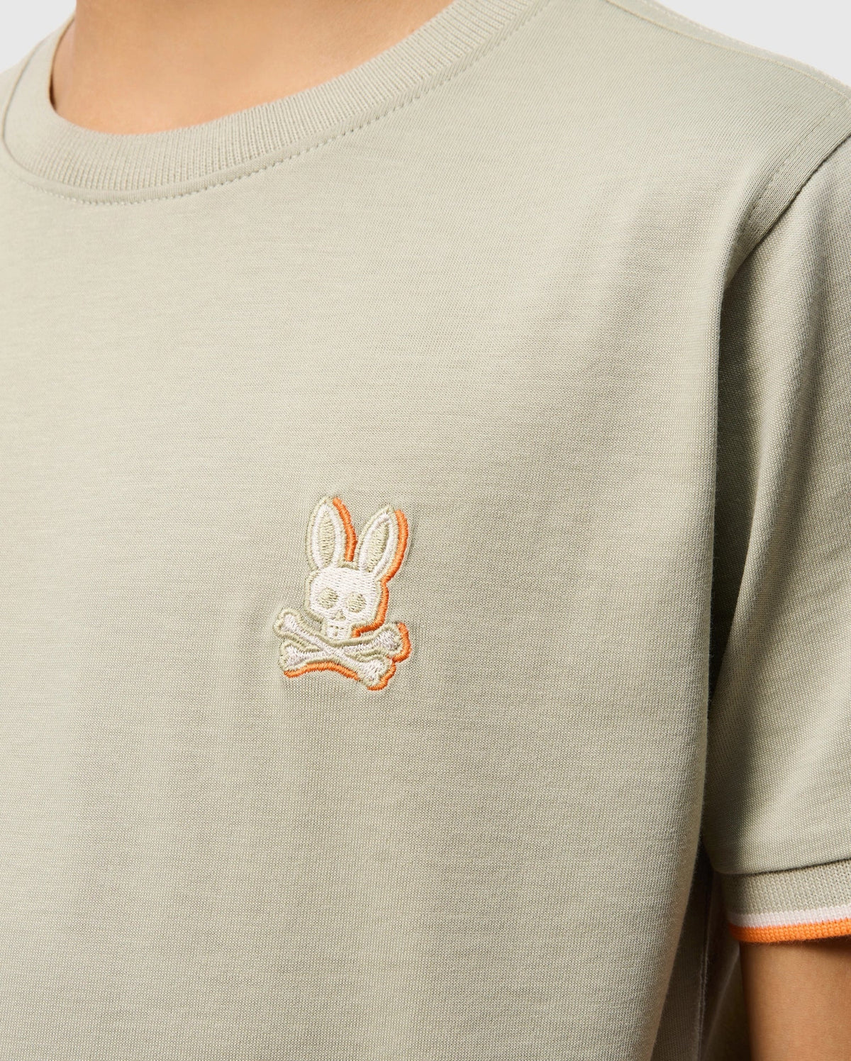 Close-up of a person wearing a light grey KIDS KAYDEN FASHION TEE - B0U577C200 from Psycho Bunny, crafted from Pima cotton, featuring an embroidered chest logo. The logo depicts a skull with rabbit ears above two crossed bones, outlined subtly in orange. The person's face is not visible in the image.