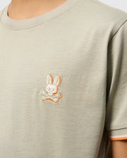 Close-up of a person wearing a light grey KIDS KAYDEN FASHION TEE - B0U577C200 from Psycho Bunny, crafted from Pima cotton, featuring an embroidered chest logo. The logo depicts a skull with rabbit ears above two crossed bones, outlined subtly in orange. The person's face is not visible in the image.