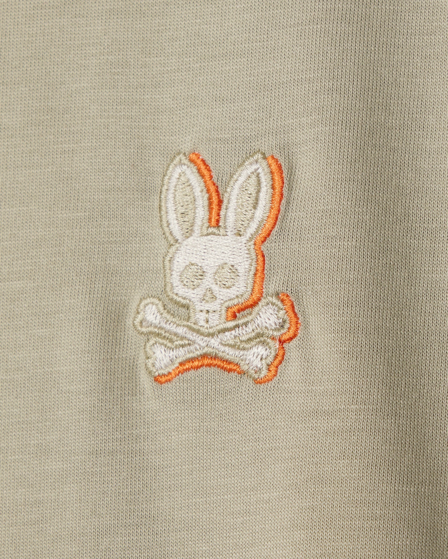 Close-up of a beige Pima cotton fabric with an embroidered chest logo featuring a stylized skull and crossbones, with the skull having bunny ears and highlighted with an orange outline. Perfect for a Psycho Bunny KIDS KAYDEN FASHION TEE - B0U577C200.