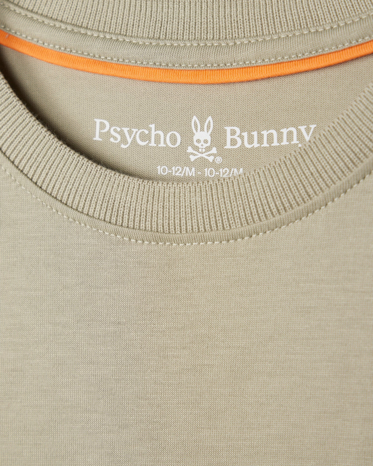 Close-up of a beige Psycho Bunny KIDS KAYDEN FASHION TEE - B0U577C200 featuring the brand's embroidered chest Bunny logo with crossbones, along with the size designation "10-12/M" printed on the inner collar area. Orange trim is visible along the inside of the collar. Crafted from soft Pima cotton for ultimate comfort.