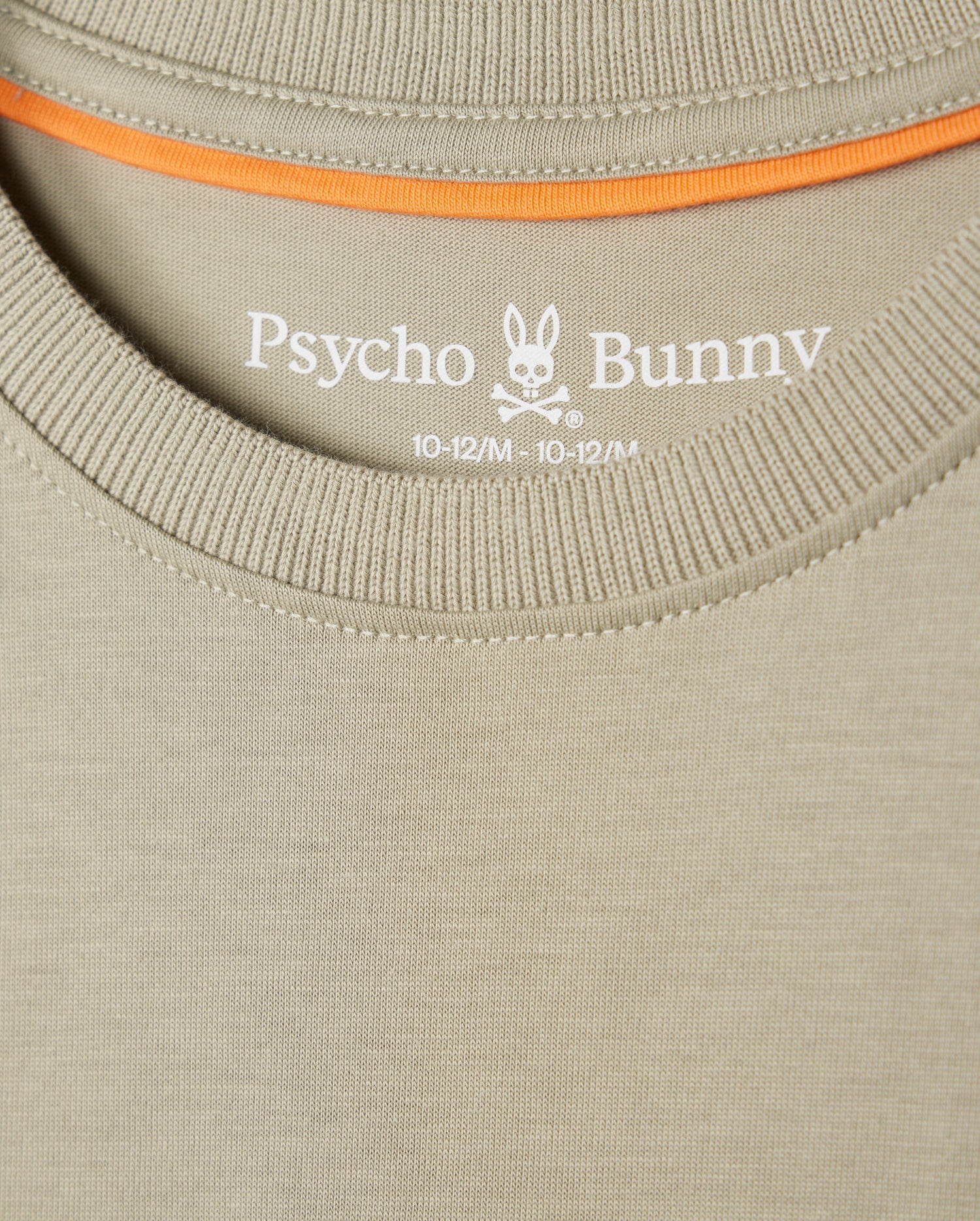 Close-up of a beige Psycho Bunny KIDS KAYDEN FASHION TEE - B0U577C200 featuring the brand's embroidered chest Bunny logo with crossbones, along with the size designation "10-12/M" printed on the inner collar area. Orange trim is visible along the inside of the collar. Crafted from soft Pima cotton for ultimate comfort.