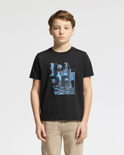 A boy stands against a plain background wearing the Psycho Bunny KIDS ZEALAND GRAPHIC TEE - B0U604F200 in black, made from soft Pima cotton, with beige pants. The shirt features a stylized blue and white pattern with abstract elements and a playful bunny logo.