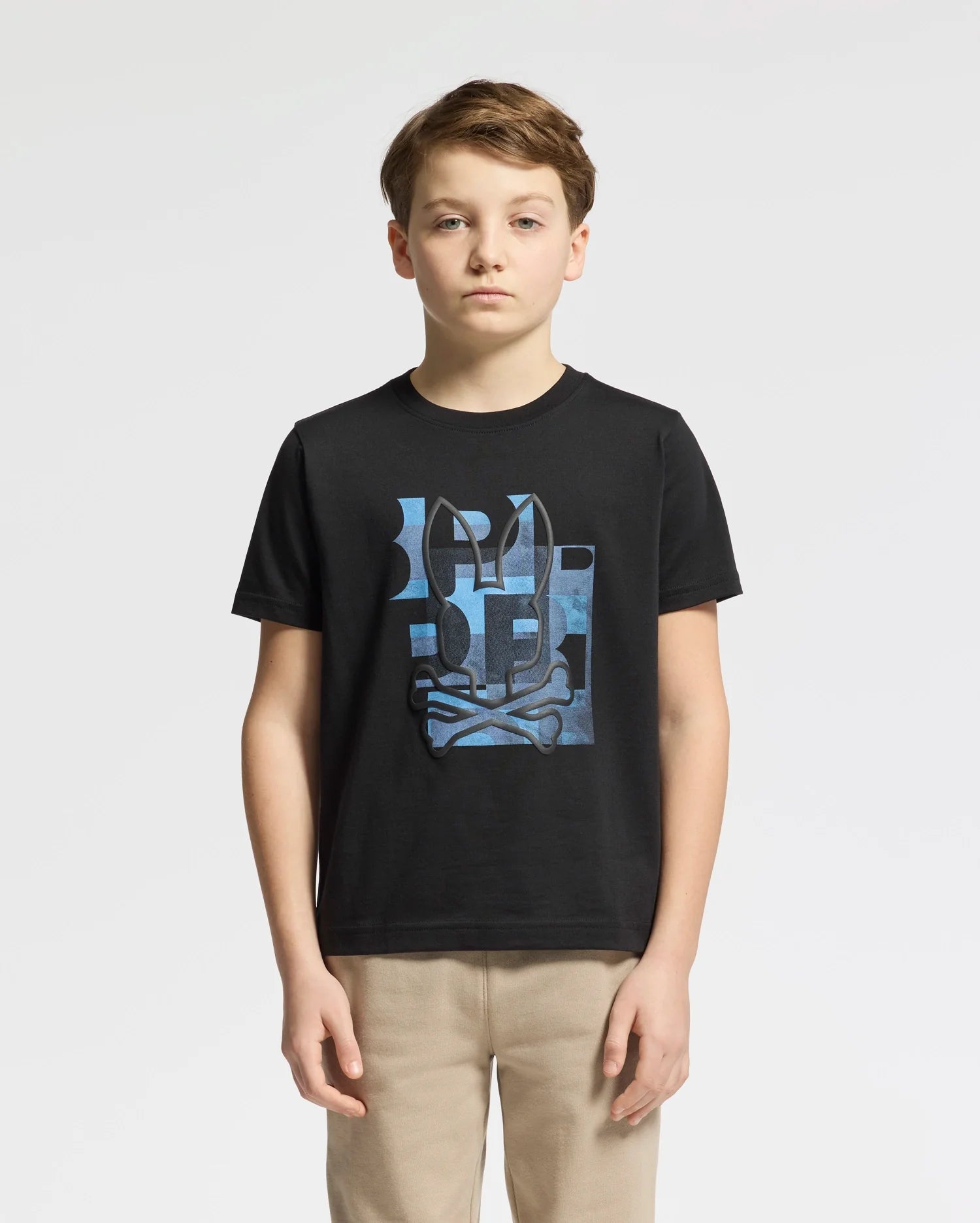 A boy stands against a plain background wearing the Psycho Bunny KIDS ZEALAND GRAPHIC TEE - B0U604F200 in black, made from soft Pima cotton, with beige pants. The shirt features a stylized blue and white pattern with abstract elements and a playful bunny logo.