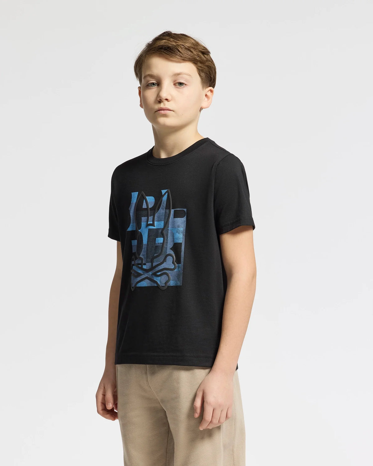 A young boy stands against a plain background, wearing a Psycho Bunny KIDS ZEALAND GRAPHIC TEE in black with a bunny logo and beige pants. He looks directly at the camera with a neutral expression.